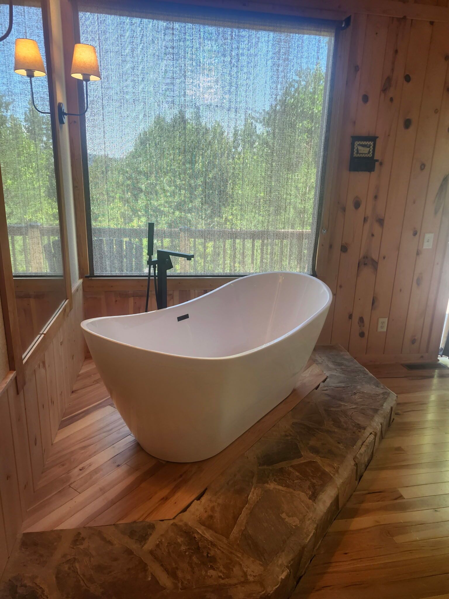 A modern bathroom with a freestanding bathtub, wooden walls, and a large window providing a view of greenery outside.