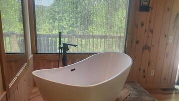 A modern bathroom with a freestanding bathtub, wooden walls, and a large window providing a view of greenery outside.