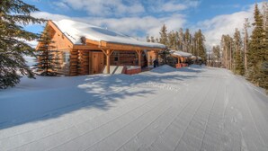 Eine Blockhütte mit schneebedecktem Dach, umgeben von hohen Kiefern und blauem Himmel.