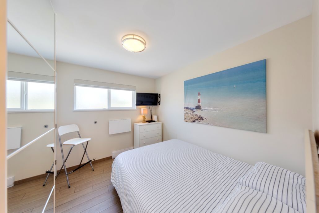 A bedroom with a bed, a chair, a desk, a flat-screen TV, a painting of a lighthouse, and a window.