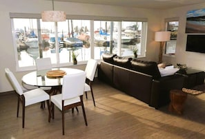 A living room with a glass table, white chairs, a brown leather sofa, a lamp, and a view of boats through large windows.