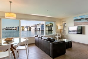 A modern living room with a glass table, white chairs, a black sofa, a TV mounted on the wall, and a large window overlooking a waterfront.