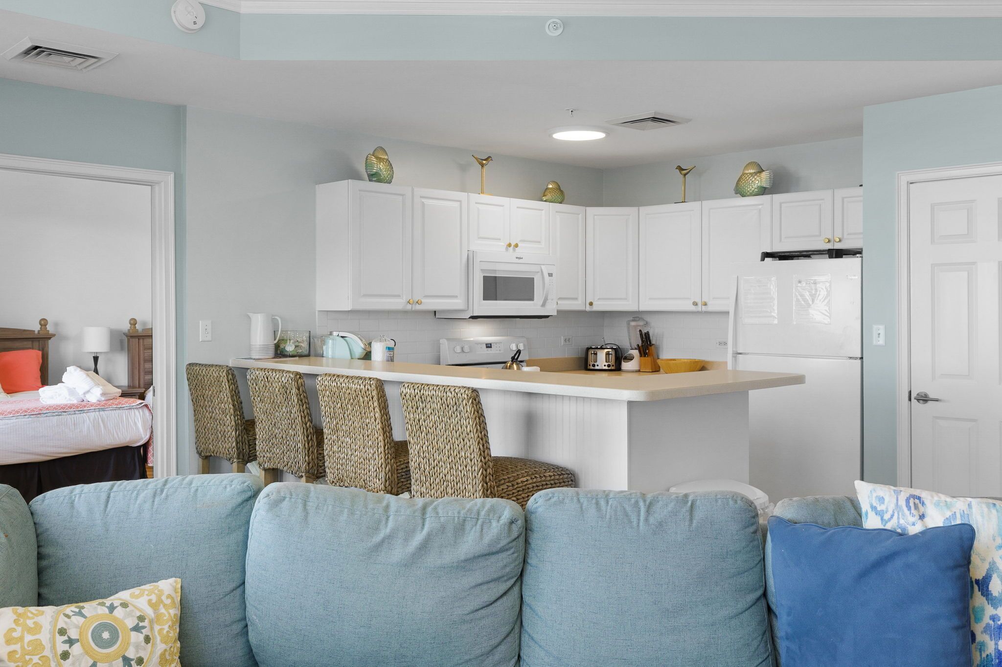 Cocina compacta con armarios blancos, microondas y refrigerador. Hay una pequeña isla con tostadora y hervidor. La habitación tiene una pared azul claro y un sofá color beige con cojines estampados.