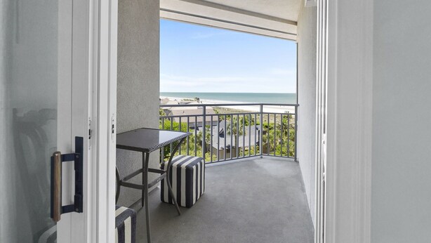 A balcony with a table and a striped ottoman, overlooking a beach and greenery. - Stunning Ocean Views from this Beautiful Condo with Pool, On-Site Amenities! (Hilton Head)