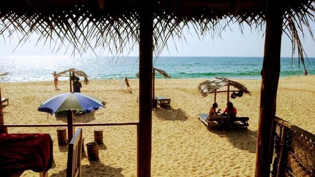 A beach view from a wooden structure with a thatched roof, featuring a sandy beach, a small table, and a chair. - Malika's Yellow House (Hikkaduwa)