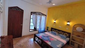 A bedroom with a bed, a wooden wardrobe, a window with curtains, a wall-mounted shelf, and a ceiling fan. - Malika's Yellow House (Hikkaduwa)