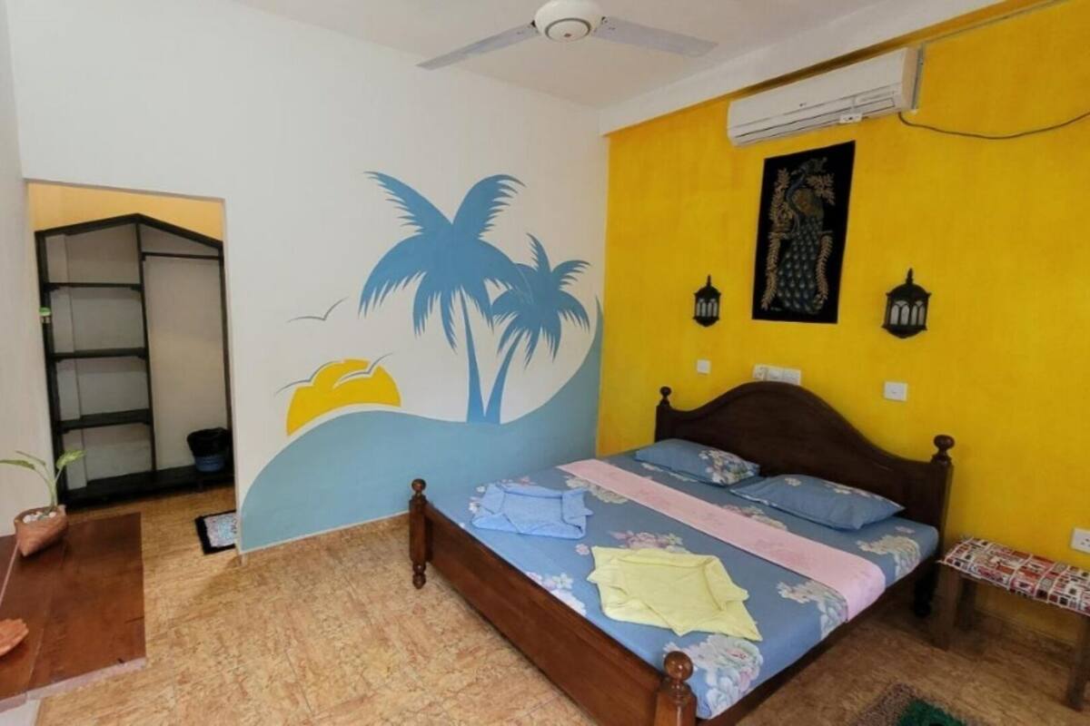 Ein Schlafzimmer mit Bett, Deckenventilator, Klimaanlage, einem Regal und einer Wand mit Palmenmotiv.