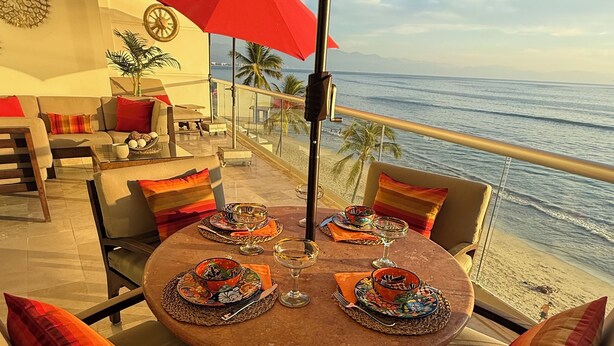 A round table with a red umbrella, surrounded by cushioned seating with colorful pillows, set on a terrace with a view of the beach and ocean. - Beautifully decorated Beachfront/Oceanfront Luxury Condo in Bucerias  (Bucerias)
