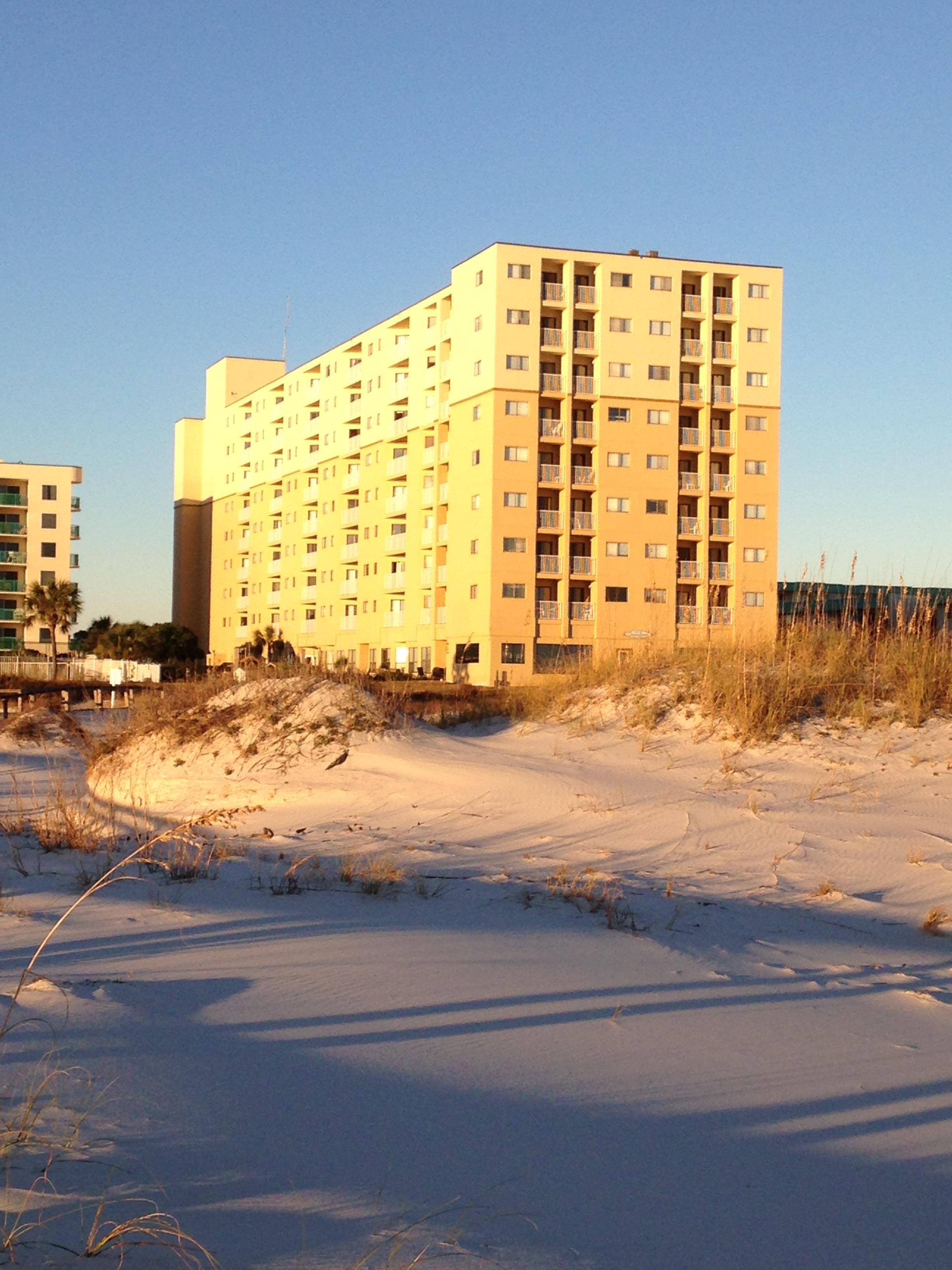 A multi-story hotel with a flat roof, located on a sandy beach with sparse vegetation.