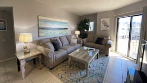 Un salon avec un canapé beige, une table basse en verre, un tapis à motifs et une grande fenêtre donnant sur des immeubles.