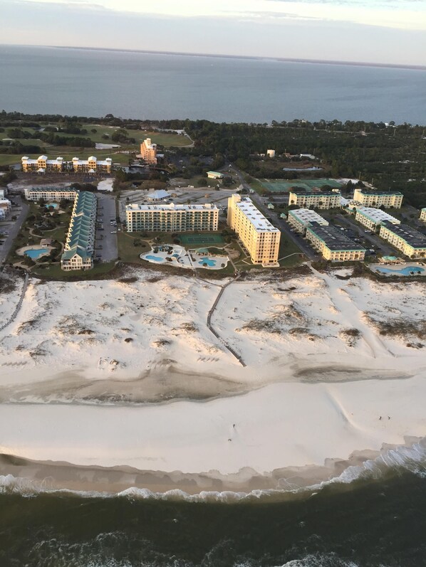 Vista aérea de un complejo turístico costero con varios edificios de hotel, una playa de arena y una extensa masa de agua al fondo.
