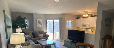 A living room with a sofa, coffee table, and chairs. There is a television, a kitchen area with cabinets, and a dining table with woven placemats.