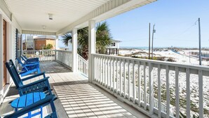 A covered porch with blue chairs, a view of the beach, and a building in the background. - Anne’s Island Girl 🌊 Gulf View Home w/ Private Pool | Tiki Bar | Pet Friendly (Pensacola Beach)