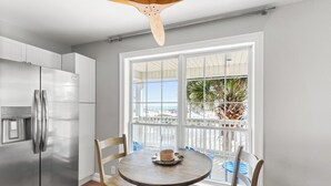 A kitchen with a stainless steel refrigerator, a wooden dining table with chairs, and a large window with a view of the beach. - Anne’s Island Girl 🌊 Gulf View Home w/ Private Pool | Tiki Bar | Pet Friendly (Pensacola Beach)
