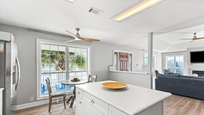A modern kitchen with a central island, white cabinetry, and a large window offering a view of palm trees. - Anne’s Island Girl 🌊 Gulf View Home w/ Private Pool | Tiki Bar | Pet Friendly (Pensacola Beach)
