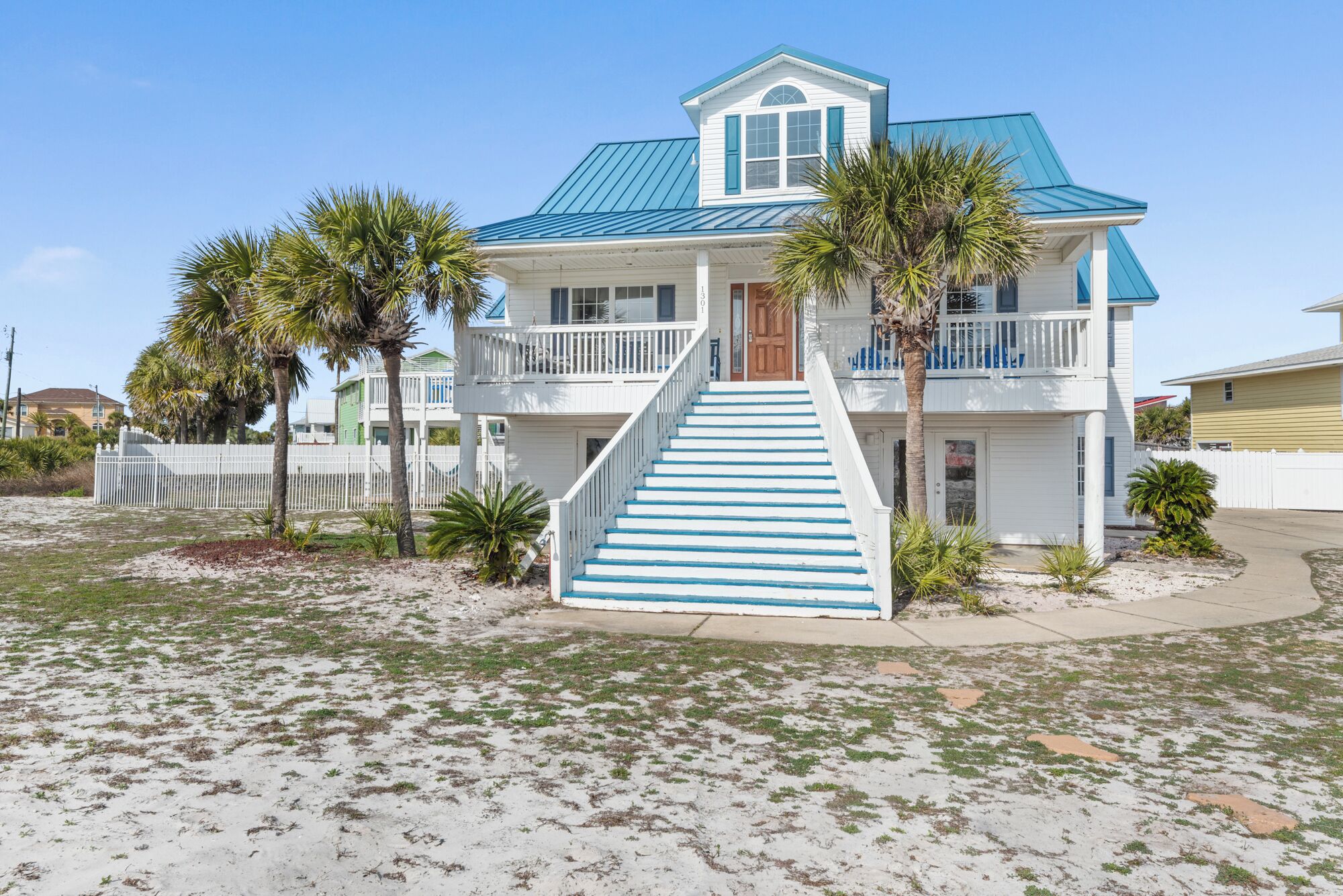 A two-story beach house with a blue roof and white exterior, featuring a front porch and a staircase leading to the entrance.