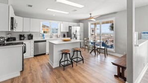 A modern kitchen with white cabinetry, a central island with stools, and a view of the beach through large windows. - Anne’s Island Girl 🌊 Gulf View Home w/ Private Pool | Tiki Bar | Pet Friendly (Pensacola Beach)