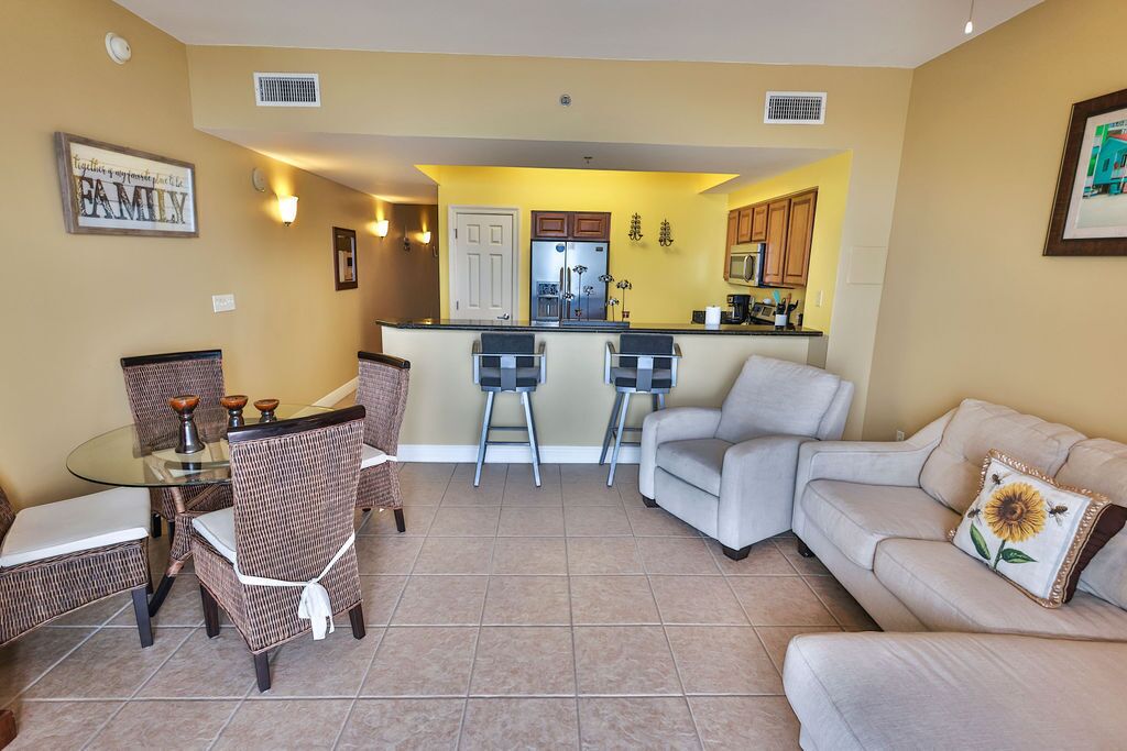 A living room with a glass dining table, wicker chairs, a sofa, and a kitchen area with a refrigerator and microwave.