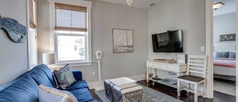 A living room with a blue sofa, a white desk, a television, and a window with blinds.