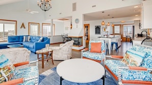 Un salon spacieux avec un canapé bleu, un fauteuil blanc et une table basse ronde. On aperçoit au fond une cheminée, un coin repas et une cuisine.