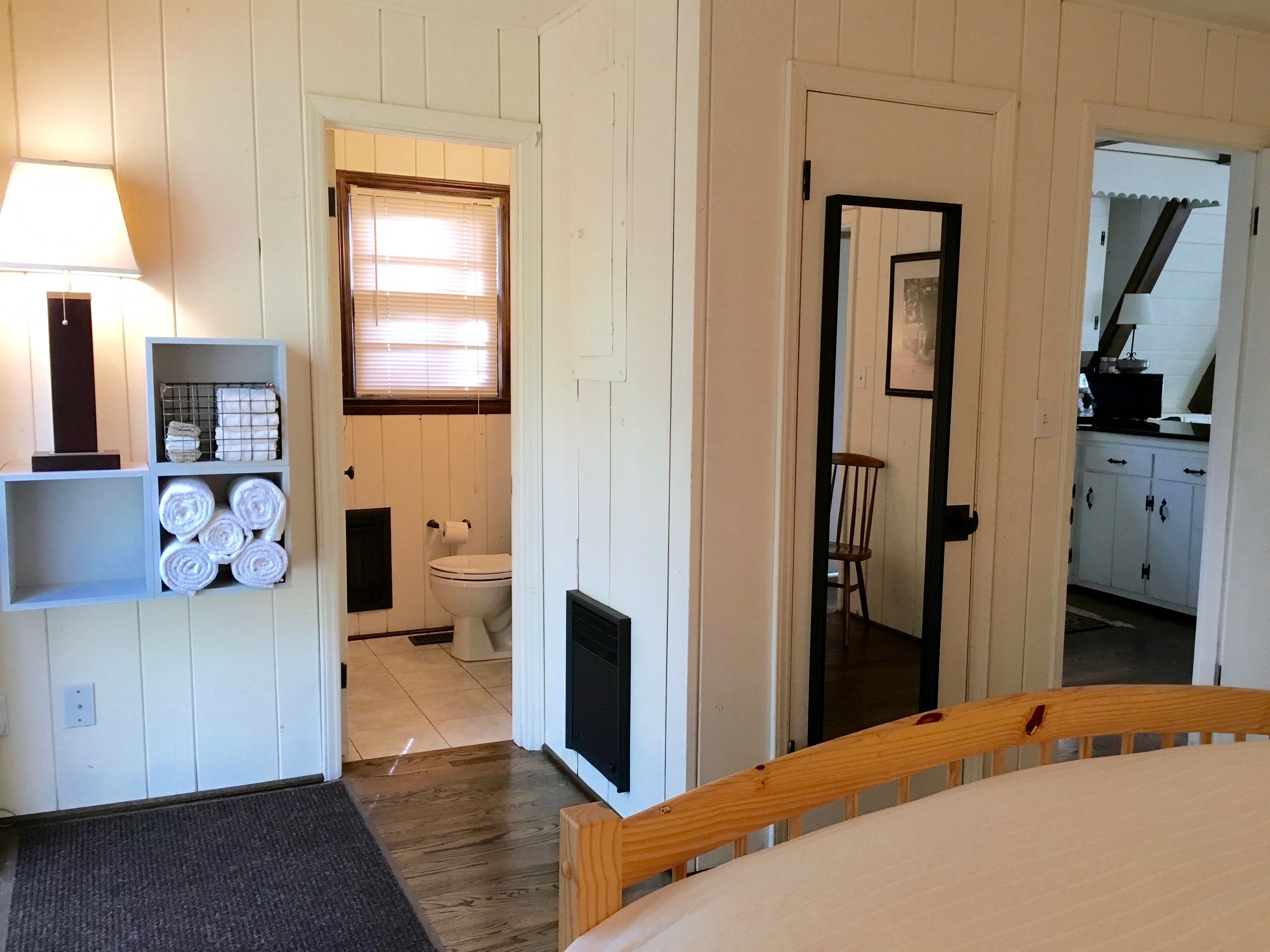 A bedroom with a bed, a nightstand, a lamp, a shelf with towels, a window with blinds, a door leading to a bathroom, and a chair.
