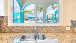 Cocina con pileta doble, encimera de granito y vista a un área de piscina con palmeras y cielo despejado.