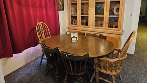 Ein Essbereich mit einem Holztisch und Stühlen, einem roten Vorhang und einer Holztruhe mit Glastüren, die Bücher und Dekorationsobjekte beherbergt.