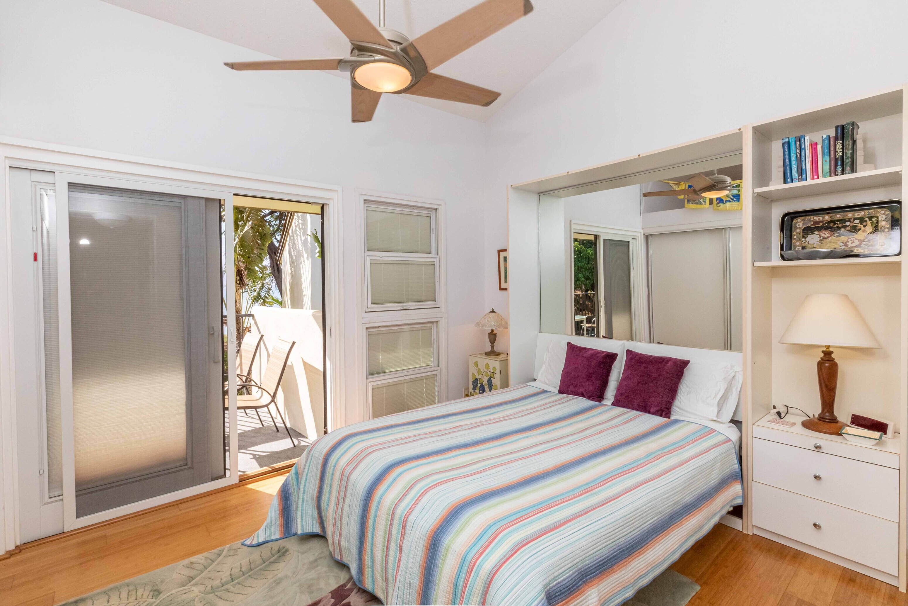 Un dormitorio con cama, ventilador de techo, mesita de noche, estantería y puerta corrediza de vidrio que da a un espacio exterior.