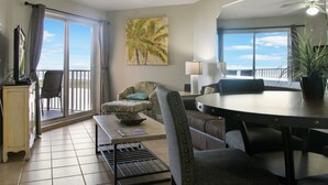 A living room with a balcony, a dining area, and a TV on a cabinet. - Surf Beach 502 - Spectacular Sunsets - Beach Front (Treasure Island)