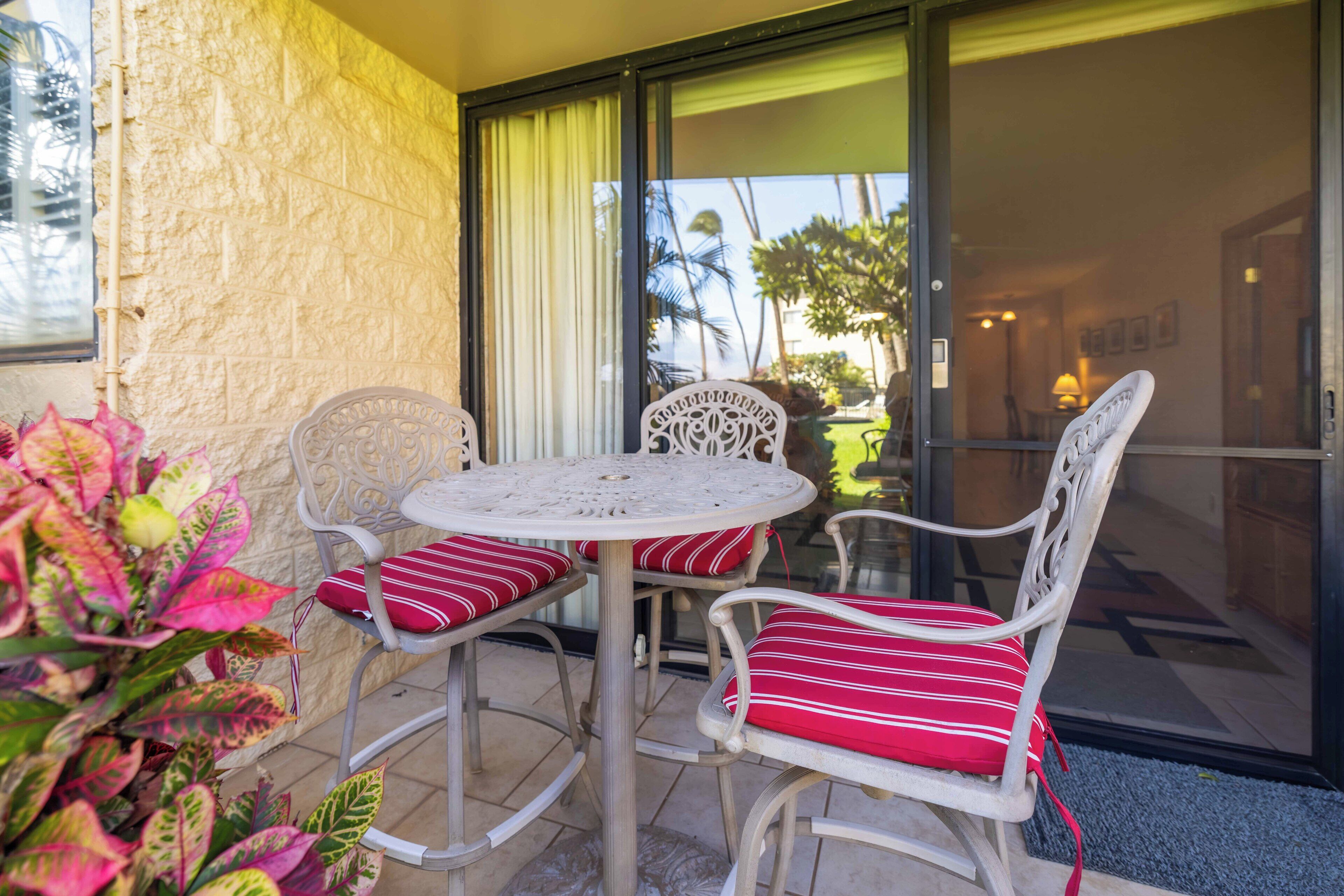 An outdoor patio with a round table and two white chairs with red cushions, a stone wall, and a glass door leading to a view of palm trees.