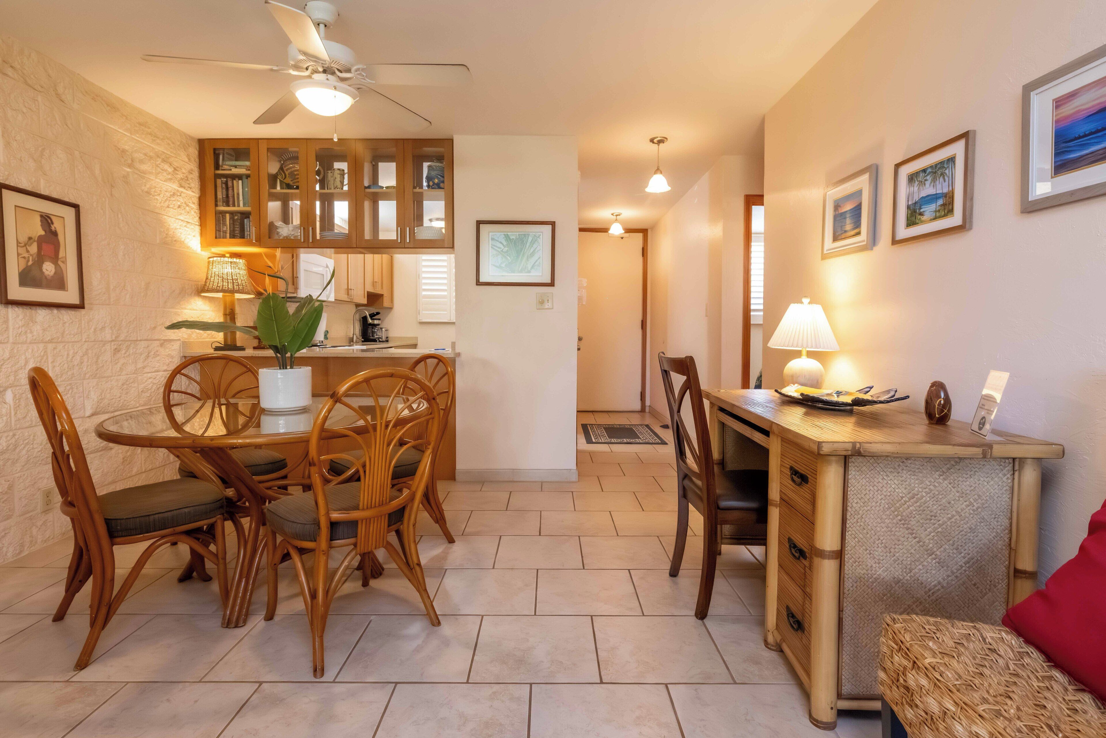 A dining area with a round table and chairs, a kitchen with wooden cabinets, a ceiling fan, and a small side table with a lamp.