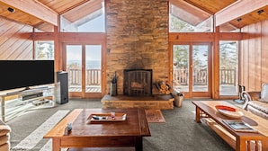 A spacious living room with a stone fireplace, wooden ceiling, and large windows offering a view of the outdoors. - SLOPESIDE CHALET, Near the top of Shay's Revenge / Ski in-Ski out / Great Views  (Snowshoe)