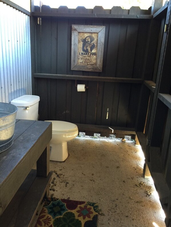 A small, enclosed outdoor bathroom with a wooden exterior, a toilet, a bench, and a framed poster on the wall. - Waterfall Pool, Cabana, Mountain Views, Outdoor Fireplace, 3 Minutes 2 Frio! (Concan)