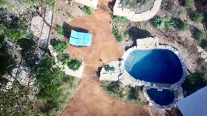 A large outdoor swimming pool surrounded by a stone wall, with a small patio area and a few lounge chairs. - Waterfall Pool, Cabana, Mountain Views, Outdoor Fireplace, 3 Minutes 2 Frio! (Concan)