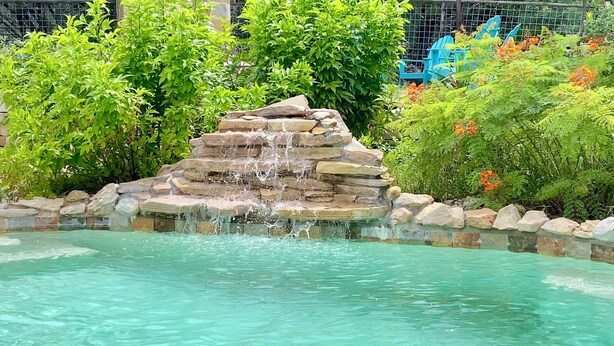A clear pool with a stone waterfall feature surrounded by lush greenery and a metal fence. - Waterfall Pool, Cabana, Mountain Views, Outdoor Fireplace, 3 Minutes 2 Frio! (Concan)