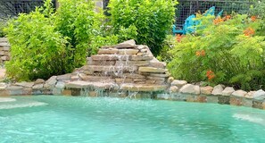 A clear pool with a stone waterfall feature surrounded by lush greenery and a metal fence. - Waterfall Pool, Cabana, Mountain Views, Outdoor Fireplace, 3 Minutes 2 Frio! (Concan)
