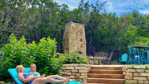 A patio area with a stone wall, a stone chimney, and a blue outdoor seating area with chairs and a table. - Waterfall Pool, Cabana, Mountain Views, Outdoor Fireplace, 3 Minutes 2 Frio! (Concan)