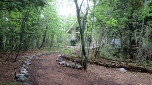 Une zone boisée avec un sentier de terre menant à un chalet. On y trouve un foyer avec des pierres disposées autour.