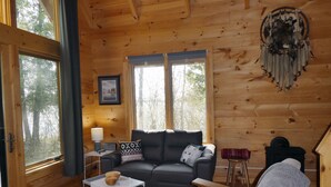 L’intérieur d’un chalet accueillant, avec un plancher en bois, un canapé gris, une petite table blanche et une grande fenêtre donnant sur la forêt.