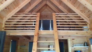 Un grenier en bois avec un plafond en pente et une fenêtre centrale. On y trouve des poutres en bois et une échelle menant à la fenêtre.