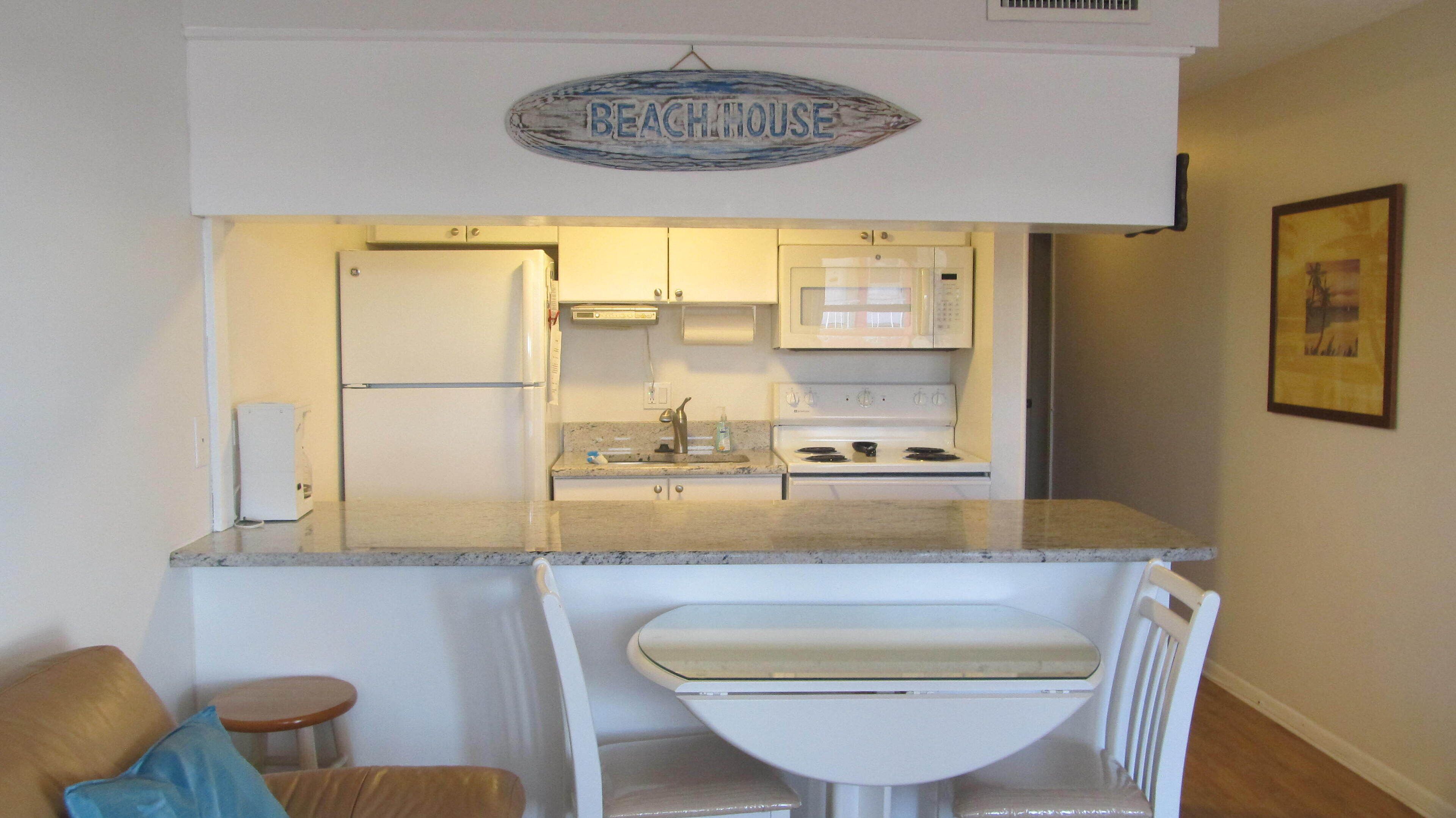 A compact kitchen with a refrigerator, microwave, stove, and a sink. A small dining area with a chair and table is visible.