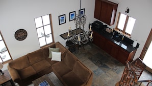 Ein Wohnzimmer mit braunem Sofa, einem Couchtisch und einer Deckenleuchte. Es gibt eine Küchenzeile mit Spüle und Schränken.