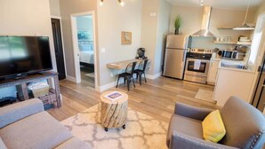 A modern living room with a flat-screen TV, a sofa, and a coffee table. The kitchen area includes a refrigerator, stove, microwave, and a dining table with chairs.