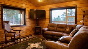 A log cabin interior with a leather sofa, a wooden chair, a fireplace, a TV, and a large window offering a view of trees. - Bitterroot Bunkhouse (Hamilton)