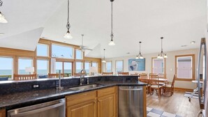 Une cuisine avec un sol à carreaux noirs et blancs, des meubles en bois, des appareils électroménagers en acier inoxydable et de grandes fenêtres offrant une vue sur l’océan.
