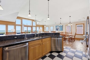 A kitchen with a black and white checkered floor, wooden cabinets, stainless steel appliances, and large windows offering a view of the ocean. - 5727- Whalebone Station, | Oceanfront | South Nags Head | Sleeps 18 (Nags Head)