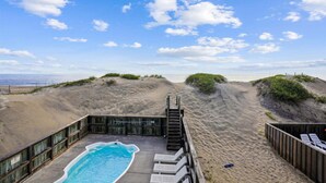 Une piscine située au bord de la plage, avec une terrasse en bois, un escalier menant à l’eau et une vue sur l’océan.