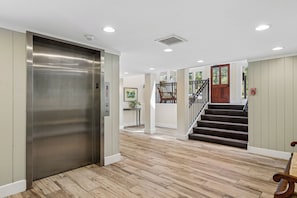 Un hall d’hôtel avec un ascenseur métallique, un escalier et un plancher en bois.