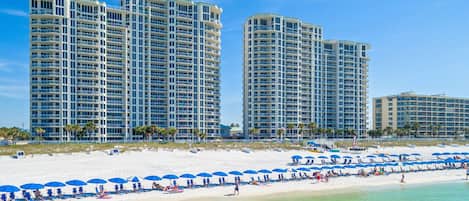 A beachfront hotel with multiple high-rise buildings, a sandy beach with blue umbrellas, and clear turquoise water.
