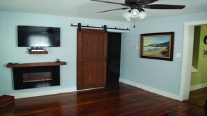 A room with a flat-screen TV mounted on the wall, a wooden cabinet below it, a sliding barn-style door, a ceiling fan, and a framed landscape painting on the wall. - Gabouri Place ~ Completely Renovated Victorian in Ste. Genevieve's Historic Dist (Ste. Genevieve)